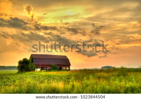American Midwest Barn Landscape. Sunset over a farm field with a traditional red barn at the horizon.

