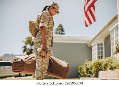 American military mom holding a teddybear while standing outside her house with her luggage. Patriotic female soldier coming back home after serving her country in the army.