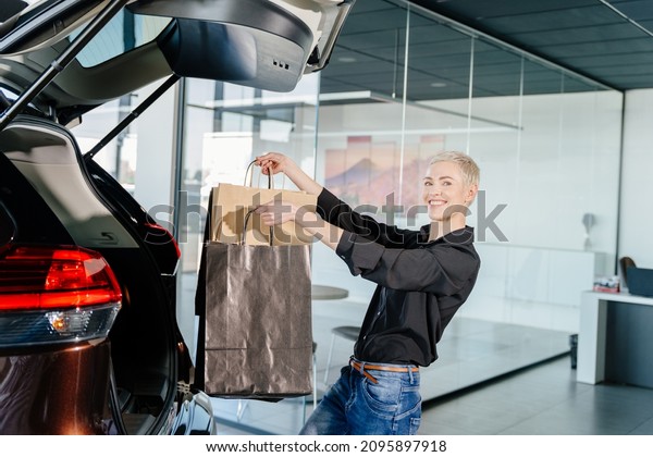 American\
short hair woman stands in car salon near vehicle with opened trunk\
and putting package in ca salon. Woman putting her shooping bags\
into car. Customer examining car at\
showroom.