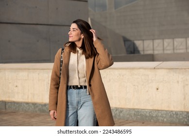 Fotografía estadounidense de una chica de ciudad tocándole el pelo, usando un abrigo de tela marrón, jeans, suéter de lana crema, pelo recto largo de hombro negro, piel muy blanca y amplia sonrisa. Fondo de los edificios grises