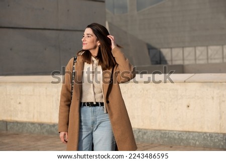 American shot of city girl touching her hair, wearing brown cloth coat, jeans, cream wool sweater, black shoulder-length straight hair, very white skin and wide smile. Background of gray buildings