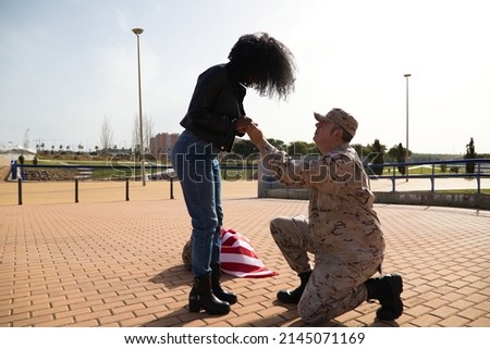 American soldier on his knees asking for marriage by holding the hands of his partner an Afro american woman and giving an engagement ring. Concept patriotism, war, soldier.