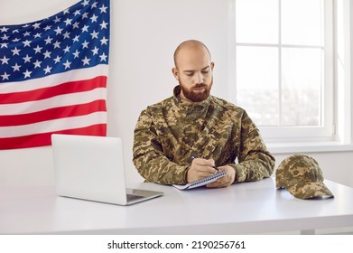 American Soldier In Uniform Sit At Desk Work On Computer Make Note In Notepad. US Military Official Use Laptop Working Online In Cabinet Write On Paper. Office Job In Army Or During War Times.