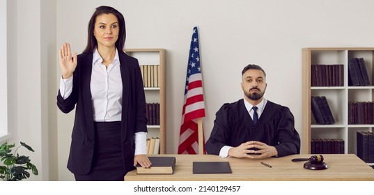 American Witness Takes An Oath Before Giving A Testimony. A Jury Member Standing Near A Male Judge In The Court Room Swears On The Bible That She Will Tell The Truth. Law And Justice In The US Concept