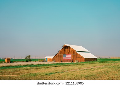 American Wooden Farm With The Flag Of United States In The Front And White Roof. Beautiful Green Farmland. 