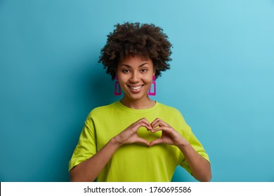 Amiable cheerful curly haired young woman makes heart shape gesture over chest, expresses love and care, wears earrings and green t shirt, isolated on blue background. Body language concept. - Shutterstock ID 1760695586