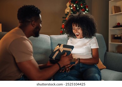 Amidst the gentle glow of the Christmas tree, an african american couple exchanges gifts with tender expressions, their shared moment radiating warmth and intimacy in a cozy setting. - Powered by Shutterstock