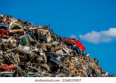 Amidst a messy heap of rusty and crumpled car parts on a junkyard, a red car sits compressed atop other scrapped vehicles, showcasing the aftermath of car wrecks and the need for waste management.