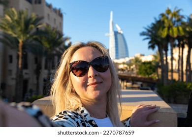 Amidst the modern architece, lively harbor, luxurious boats, towering skyscrapers, and sunny sky with clouds, a blonde tourist girl takes some selfies to remember her visit to the Dubai resort area. - Powered by Shutterstock