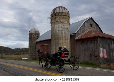 An Amish Couple Rides By In Their Buggy Pulled By A Horse With A Farm With American Flags In The Background