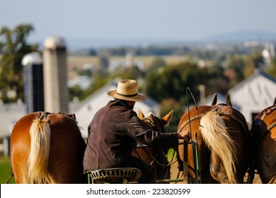 Amish Farmer On Horse Drawn Plow With Farm In Background In Rural Pennsylvania. 