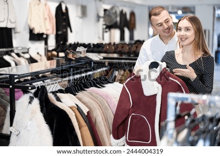 Among large clothes selection on rails in store, girl and her husband male companion stopped near showcase with outerwear, holds fur coat hanger in hands and examines product, integrity of garniture