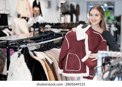 Among large selection of clothes on rails in store, girl stopped near showcase with outerwear, holds fur coat hanger in hands and examines product, integrity of garniture.