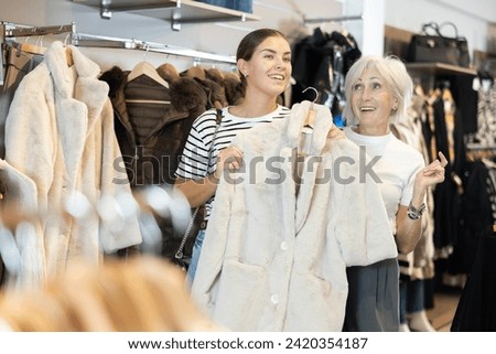 Among large selection of clothes on rails in store, girl and mature woman companion stopped near showcase with outerwear, holds fur coat hanger in hands and examines product, integrity of garniture