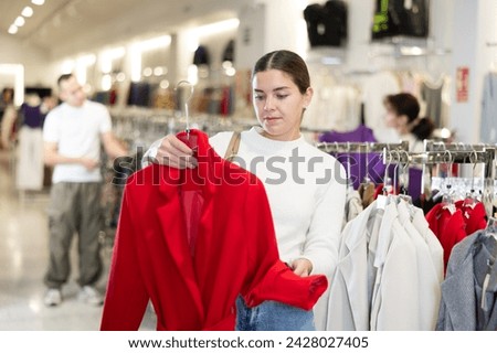 Among large selection of clothes on rails in store, girl stopped near showcase with outerwear, holds coat hanger in hands and examines product, integrity of garniture.
