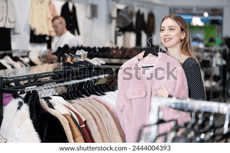 Among large selection of clothes on rails in store, girl stopped near showcase with outerwear, holds fur coat hanger in hands and examines product, integrity of garniture.