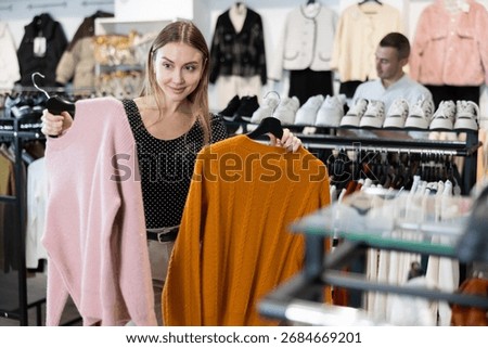 Among large selection of clothes on rails in store, girl stopped near showcase with outerwear, holds jemper hanger in hands and examines product, integrity of garniture.