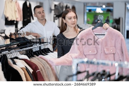 Among large selection of clothes on rails in store, girl stopped near showcase with outerwear, holds fur coat hanger in hands and examines product, integrity of garniture.