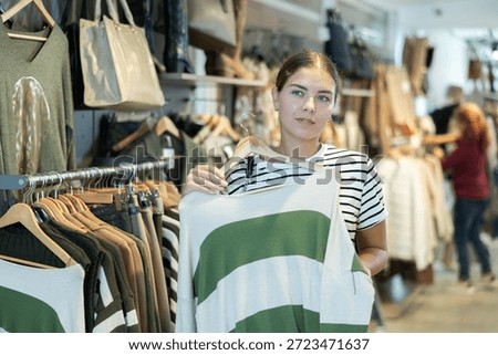 Among large selection of clothes on rails in store, girl stopped near showcase with outerwear, holds jemper hanger in hands and examines product, integrity of garniture.