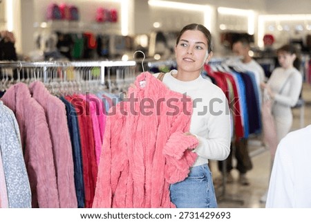 Among large selection of clothes on rails in store, girl stopped near showcase with house clothes, terrycloth robe hanger in hands and examines product, integrity of garniture.