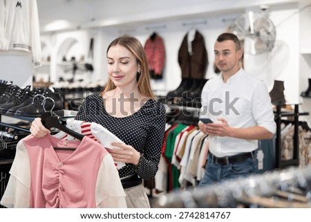 Among large selection of clothes on rails in store, girl stopped near showcase with outerwear, holds blouse hanger in hands and examines product, integrity of garniture.
