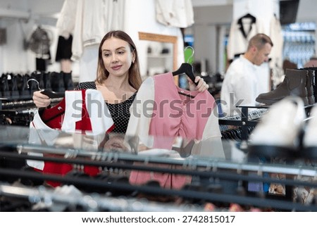 Among large selection of clothes on rails in store, girl stopped near showcase with outerwear, holds blouse hanger in hands and examines product, integrity of garniture.