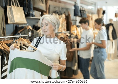 Among large selection of clothes on rails in store, senior woman stopped near showcase with outerwear, holds jemper hanger in hands and examines product, integrity of garniture.