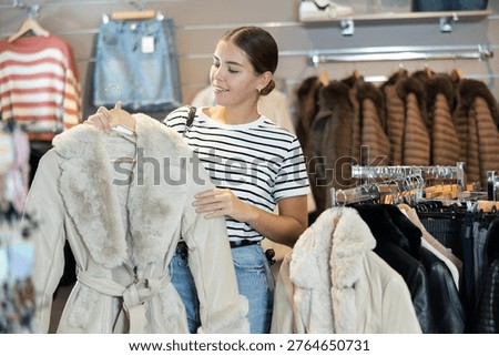 Among large selection of clothes on rails in store, girl stopped near showcase with outerwear, holds fur coat hanger in hands and examines product, integrity of garniture.