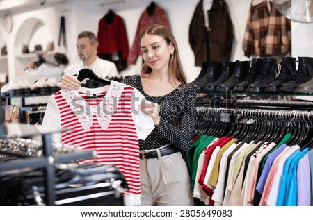 Among large selection of clothes on rails in store, girl stopped near showcase with outerwear, holds blouse hanger in hands and examines product, integrity of garniture.