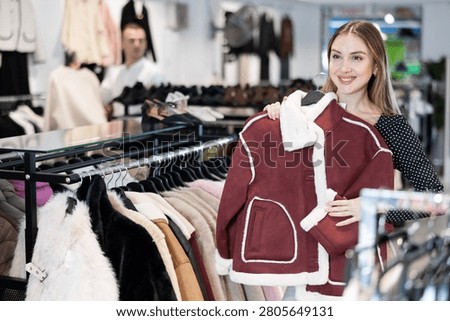 Among large selection of clothes on rails in store, girl stopped near showcase with outerwear, holds fur coat hanger in hands and examines product, integrity of garniture.