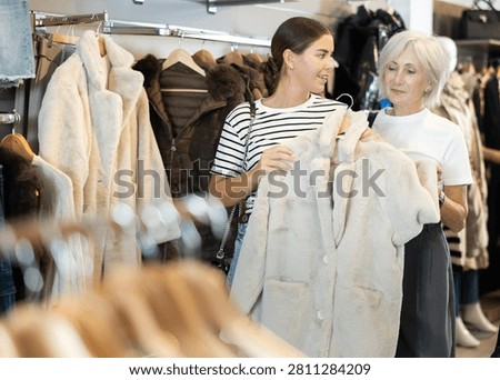 Among large selection of clothes on rails in store, girl and mature woman companion stopped near showcase with outerwear, holds fur coat hanger in hands and examines product, integrity of garniture