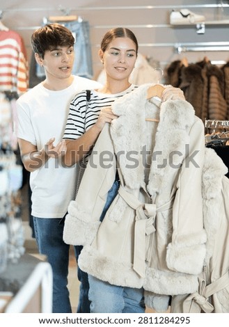 Among large selection of clothes on rails in store, girl and guy companion stopped near showcase with outerwear, holds fur coat hanger in hands and examines product, integrity of garniture