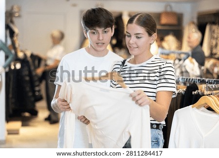Among large selection of clothes on rails in store, girl and her guy companion stopped near showcase with outerwear, holds jemper hanger in hands and examines product, integrity of garniture.