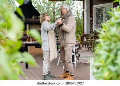 Amorous senior couple in warm casualwear dancing on wooden floor of patio by their country house against served table and fireplace
