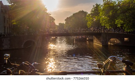 Amsterdam canal with trees, bridge and boats - the capital in Netherlands, the sun is shining and reflected in the water, the water is in front of the image and in the background is a bridge and trees