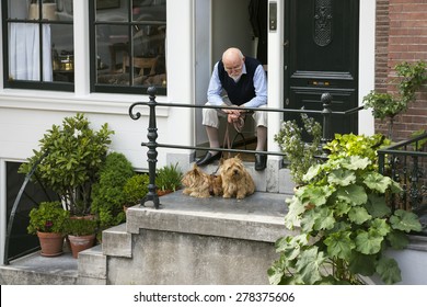 Amsterdam, Netherlands, 10 May 2015: Elderly Man Sits With Two Dogs In Doorway Of Canal House In Amsterdam On Sunny Day