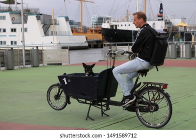 Amsterdam, Netherlands. About The July 2019.  Man And Black Dog On A Bicycle Await The Ferry.