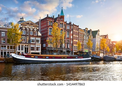 Amsterdam, Netherlands. Floating Houses and houseboats and boats at channels by banks. Traditional dutch dancing houses among trees. Evening autumn street above water pink sunset sky with clouds.