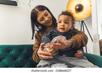 Amused 5 Years Old African American Child Sitting On His Young Caucasian Mother's Lap And Enjoying Cookies Together - Room Atmosphere Background. High Quality Photo