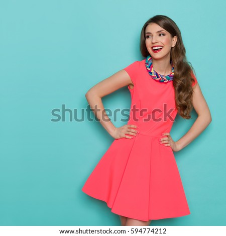 Amused beautiful young woman in pink mini dress posing with hand on hip and looking away. Three quarter length studio shot on turquoise background.