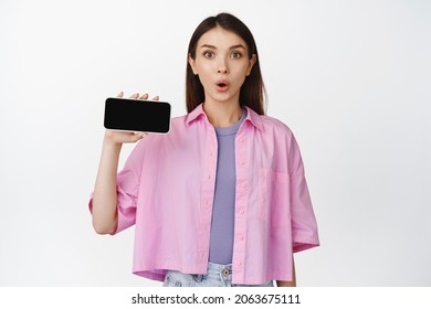 Amused Brunette Girl Gasp, Say Wow, Showing Horizontal Smartphone Screen, Standing In Casual Clothes Over White Background