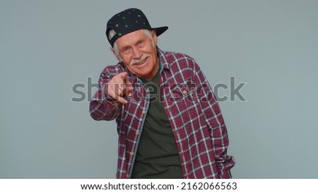 Amused senior man in casual shirt pointing finger to camera, laughing out loud taunting making fun of ridiculous appearance, funny joke. Elderly grandfather posing alone on gray studio wall background
