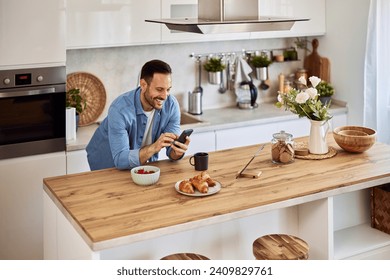 An amused young adult man laughing and scrolling through social media platforms on his smart phone while leaning on a kitchen counter in front of breakfast. - Powered by Shutterstock