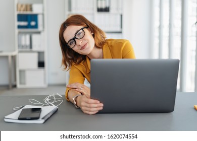 Amused Young Businesswoman Peering Around Her Laptop At The Camera With A Mischievous Smile As She Works At Her Desk In The Office