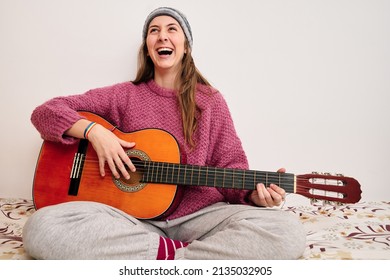 Amused Young Woman In Casual Attires Sitting Barefoot On The Bed Exclaiming With Joy After Playing A Song On Her Guitar. Hobbies And Lifestyle Concept.