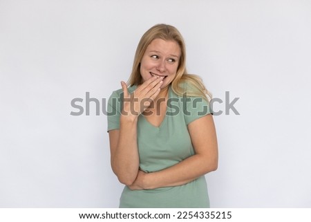 Amused young woman covering mouth. Portrait of shy Caucasian female model with fair hair in green T-shirt looking aside with hand on mouth, laughing at joke or teasing someone. Fun, happiness concept