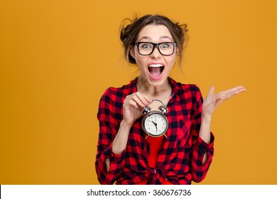 Amusing pretty young woman in plaid shirt and glasses holding alarm clock and shouting over yellow background