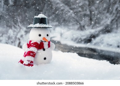 Amusing Snowman Stands In A Snowdrift With Winter River And Forest On The Background At Countryside In Cold Snowy Day