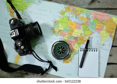 Analog camera, word map, notepad, pen and compass on wooden background