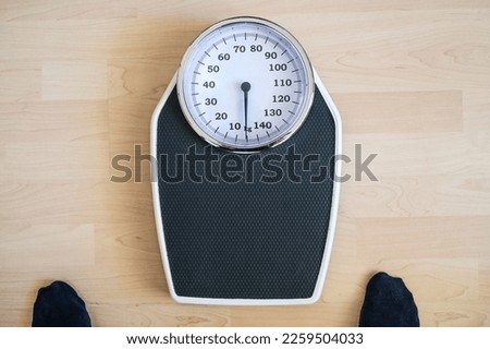 Analogue old-fashioned personal scale on a wooden floor, two feet in dark stockings hesitantly standing in front of it, worried about the weight, high angle view from above, copy space